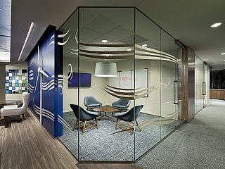 A conference room with lounge chairs behind a glass wall, another lounge grouping to the left and hallway to the right
